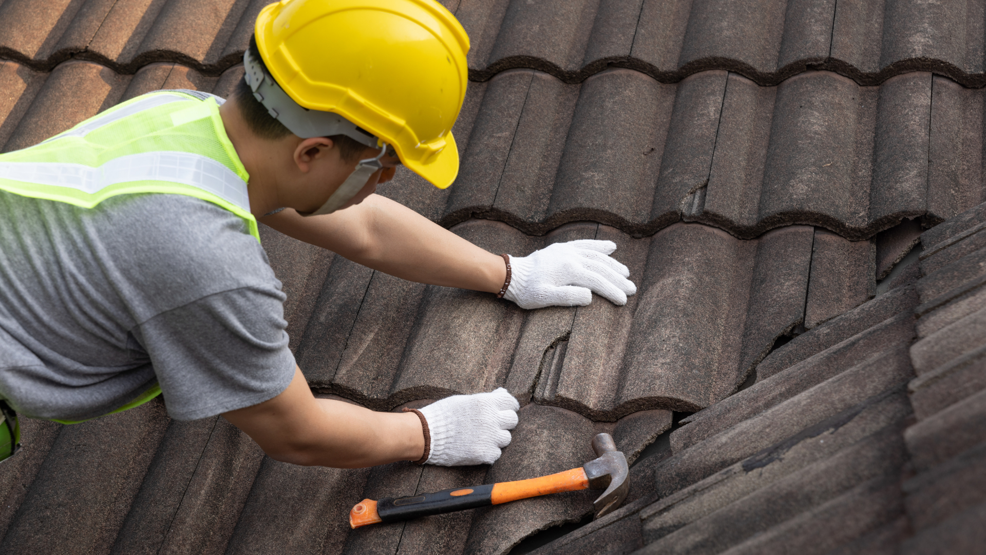 A roofing contractor wearing safety gear and a yellow hard hat inspects and repairs cracked roof tiles using a hammer during a maintenance check.