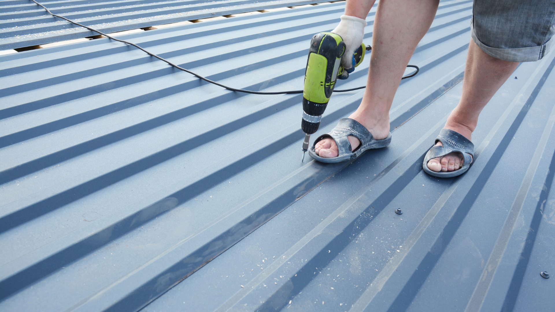A person in shorts and sandals using a power drill to screw down a dark blue corrugated metal roof panel.