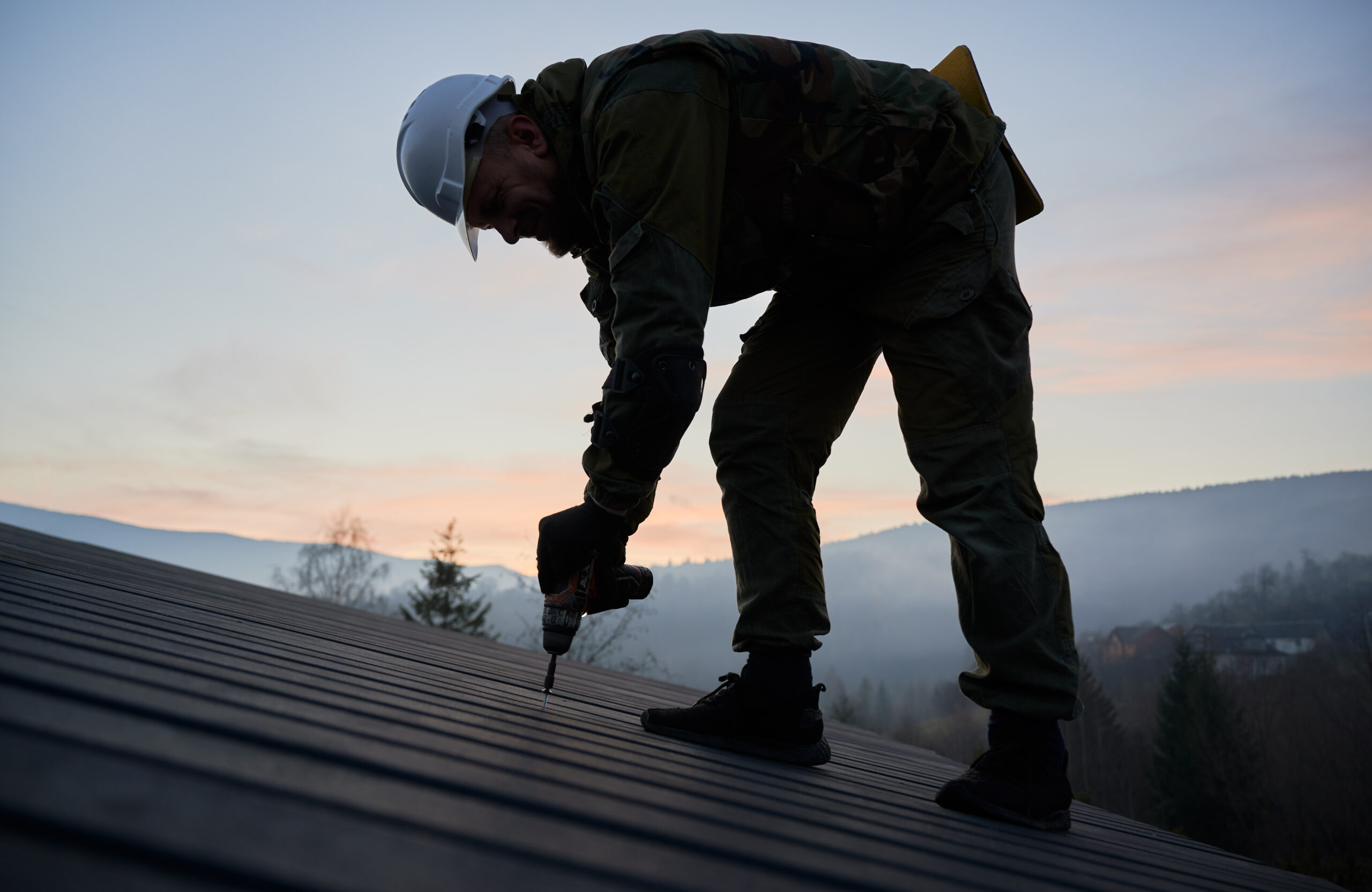 Male builder installing black corrugated iron sheet used as facade of future cottage. Silhouette of man worker building wooden frame house. Carpentry and construction concept.