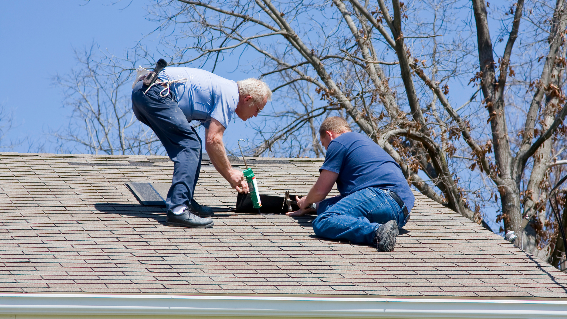 Two men repairing a residential roof with tools and sealant on a sunny day.