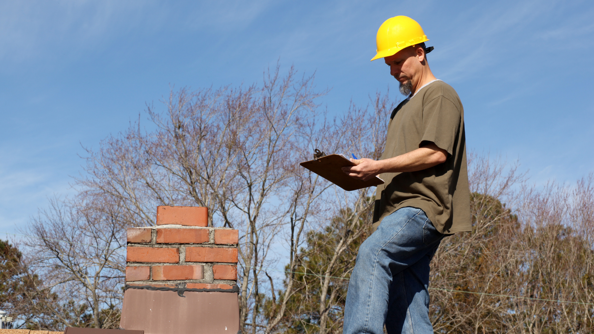 Construction worker in a yellow hard hat inspecting a brick chimney with a clipboard on a roof.