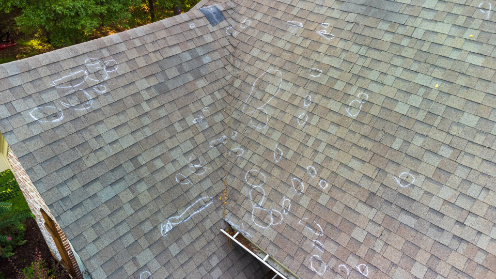 The image shows a close-up, aerial view of a residential shingle roof with numerous white chalk circles and markings. These markings highlight areas of damage, likely caused by hail. The roof has brown and gray asphalt shingles and a metal gutter is visible along the lower edge of the roof.